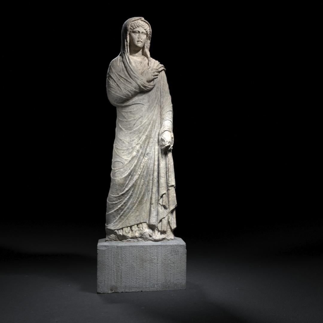 A full-length marble statue of a robed woman from ancient Rome, possibly a goddess or noblewoman, standing in a frontal pose with her head covered, illuminated against a dark background.