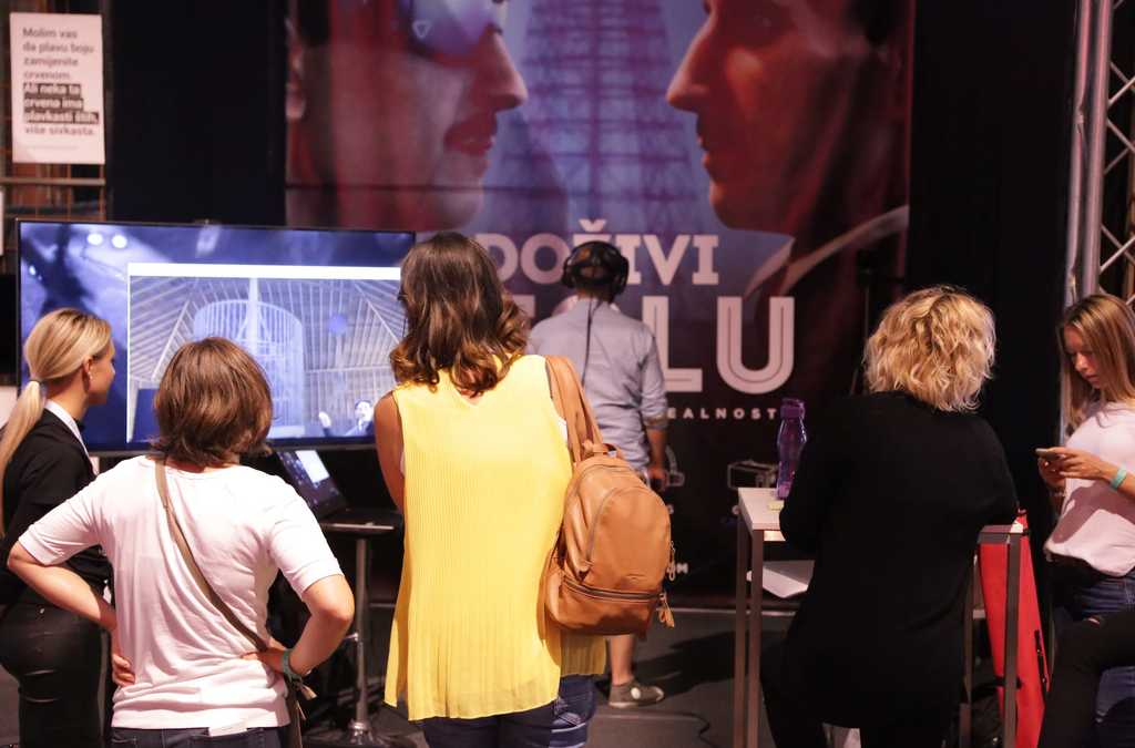 A group of people observing a person using VR equipment, with a focus on the virtual interaction displayed on a monitor amidst an exhibition setup.