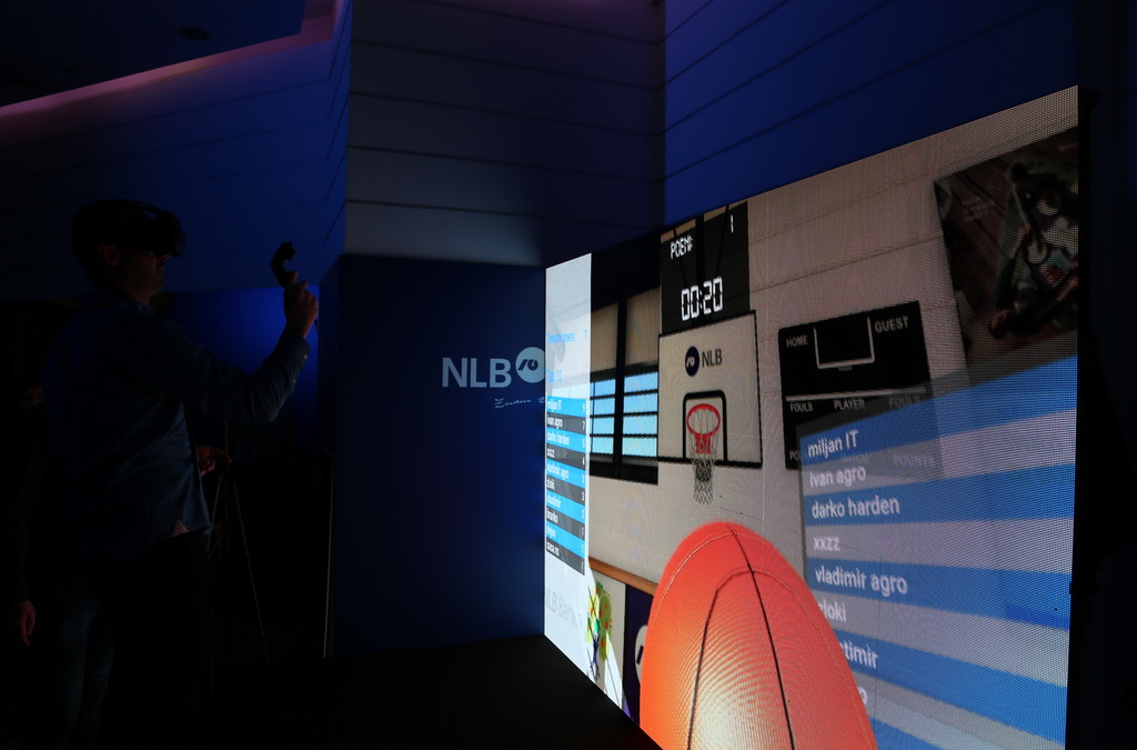Over-the-shoulder view of a VR game participant aiming with a controller at a basketball hoop on a large screen, with the game's scoreboard visible.