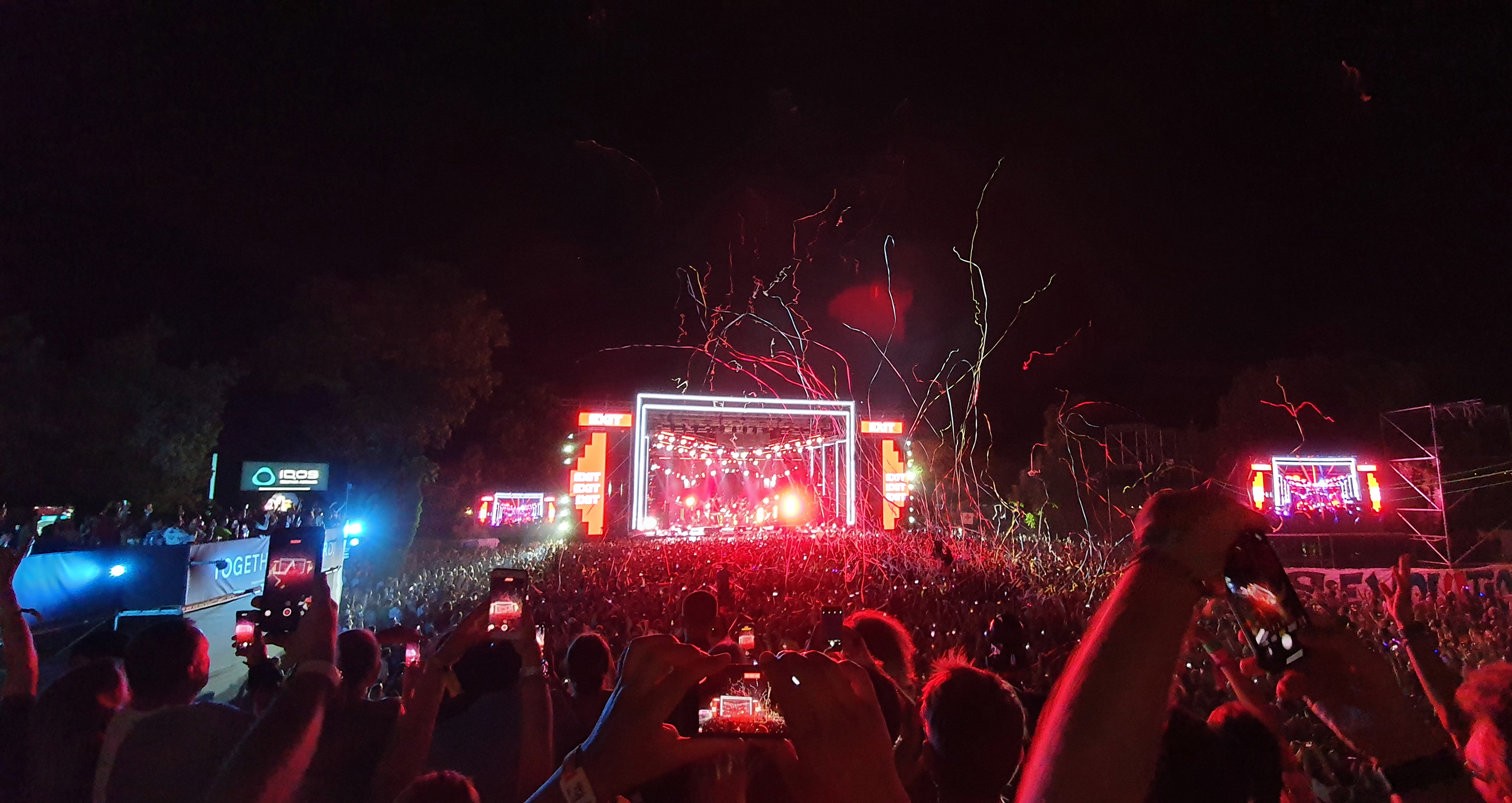 A vibrant concert scene at night, showcasing a large crowd of people gathered in front of a brightly lit stage with a musical performance in progress. Numerous hands are raised, some holding smartphones to capture the moment. 