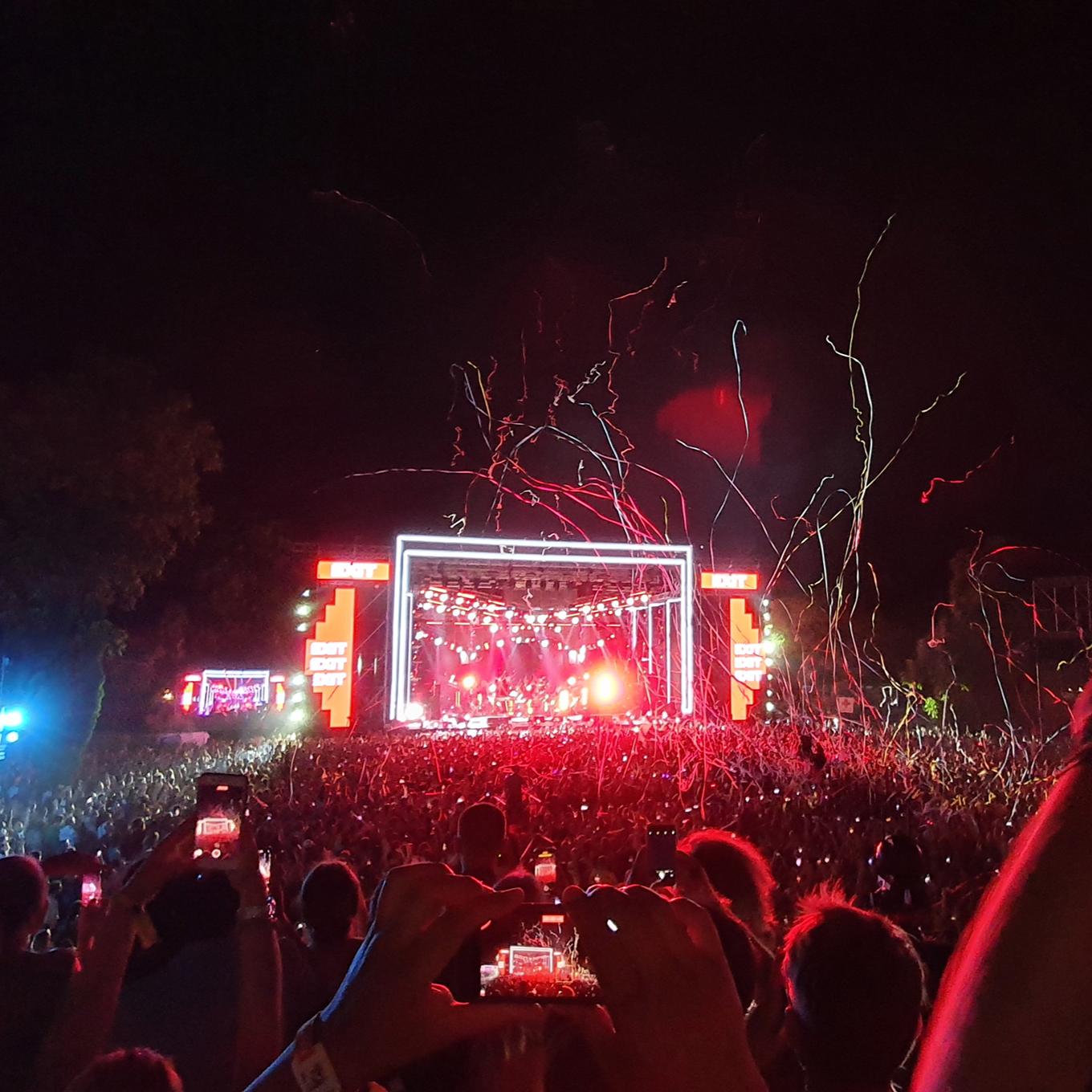 A vibrant concert scene at night, showcasing a large crowd of people gathered in front of a brightly lit stage with a musical performance in progress. Numerous hands are raised, some holding smartphones to capture the moment.