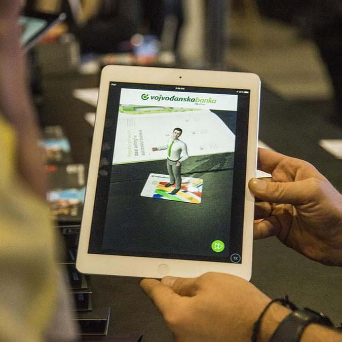 A person holds an iPad displaying Voban, Vojvođanska bank's virtual assistant, standing on a graphic of a bank card, showing off his ability to provide information interactively.