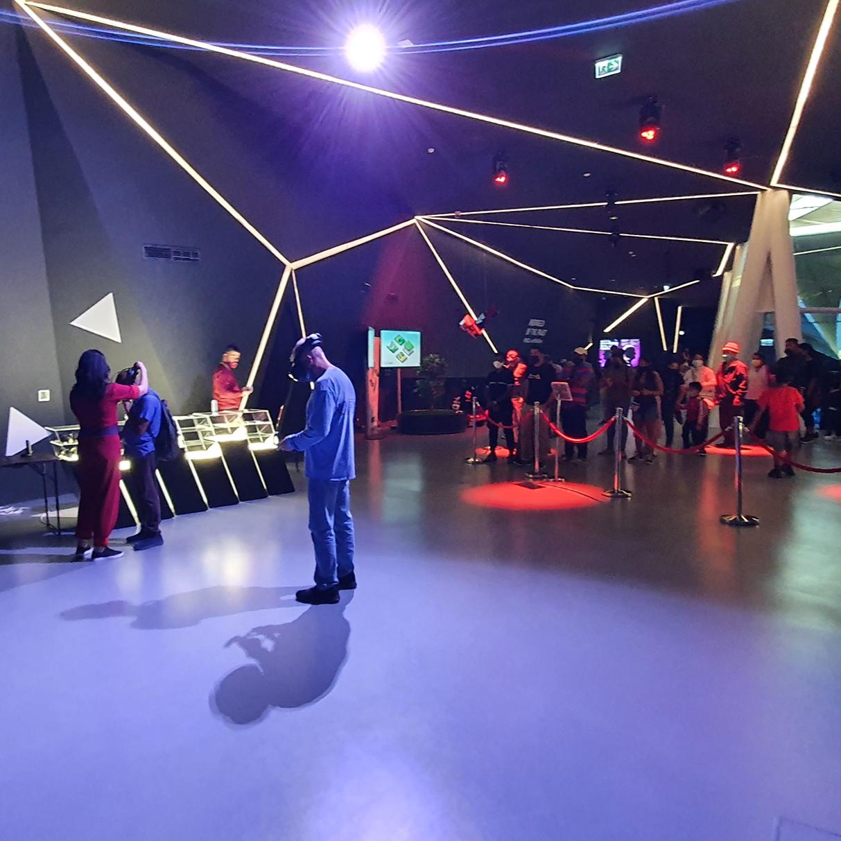 Attendees experiencing virtual reality at the Serbian Pavilion's VR Zone during Expo 2020 in Dubai. People are wearing VR headsets and interacting with the digital environment, immersed in a space illuminated by dynamic geometric light installations, reflective of Serbia's innovation in technology and arts.