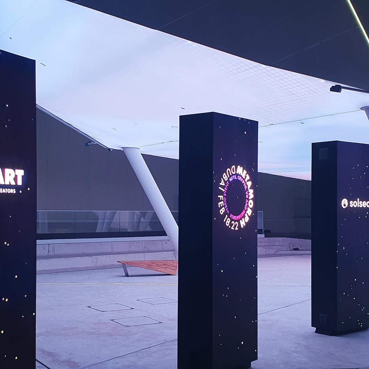 An installation features multiple illuminated pillars with branding for the ALL-ART and Solsea platforms, part of the Metamorph event by Digital Mind and VR-All-Art. This setup showcases the innovative avatar system intended to transform event experiences, highlighting the fusion of physical and virtual worlds in the context of crypto and NFTs.