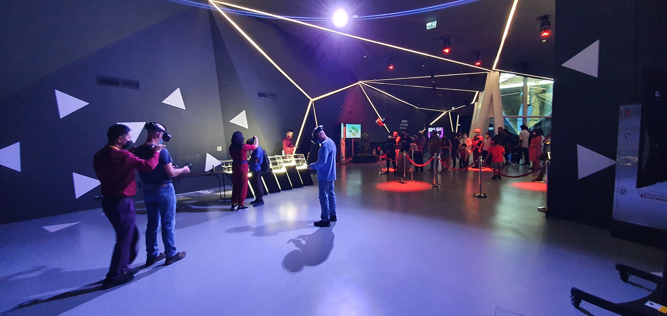 Attendees experiencing virtual reality at the Serbian Pavilion's VR Zone during Expo 2020 in Dubai. People are wearing VR headsets and interacting with the digital environment, immersed in a space illuminated by dynamic geometric light installations, reflective of Serbia's innovation in technology and arts.