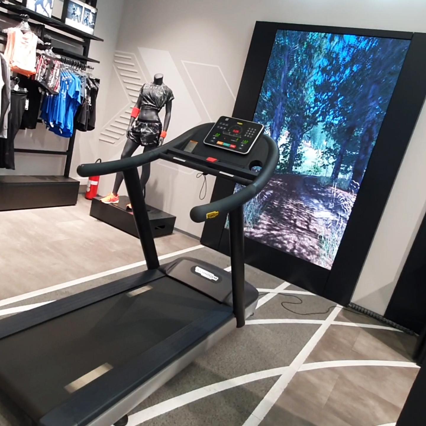 An interactive Sport Vision Running setup in a store, featuring a treadmill facing a screen with a forest simulation for a virtual outdoor running experience. The surrounding sports apparel and the store's athletic decor underscore the fusion of fitness and technology.