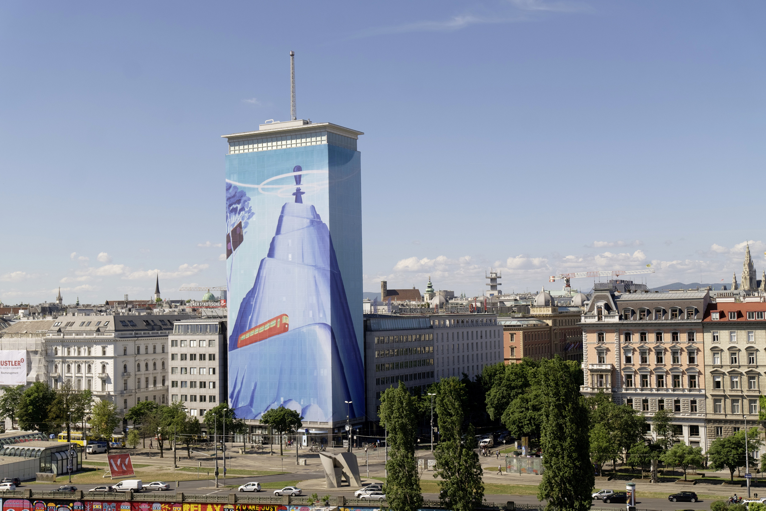 The Ringturm building in Vienna, wrapped in a colossal art installation portraying a blue-hued scene with satellite, space, and technology motifs, representing the Wiener Städtische's commitment to innovation. The surrounding urban landscape contrasts with the vibrant artwork.