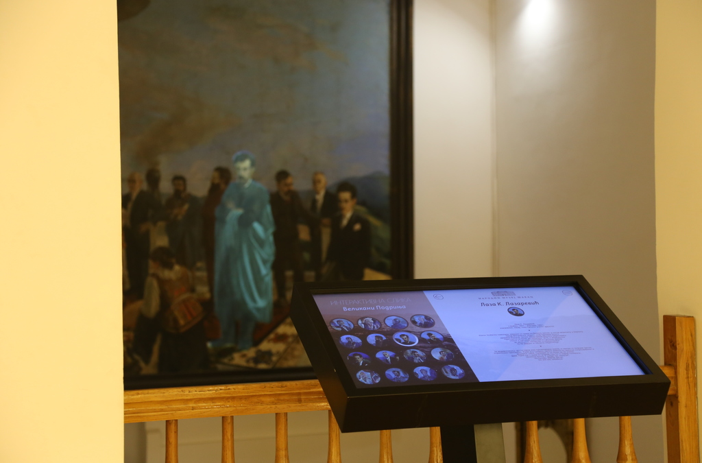 An interactive museum exhibit featuring a multi-touch screen display in front of a historical painting.