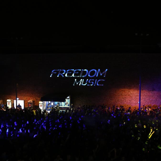 A crowd of concert-goers illuminated by neon lights, with the words "FREEDOM MUSIC" projected in large, floating letters on the brick wall above the event entrance.