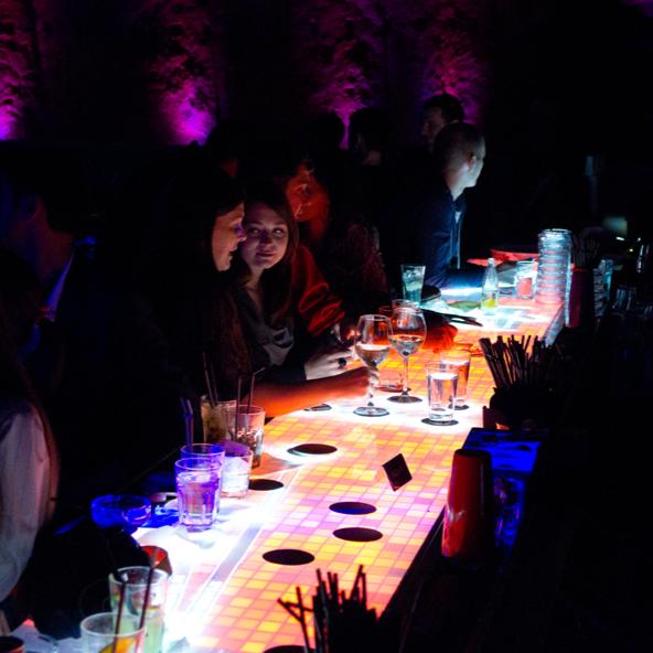 Patrons are seated at a vibrant, interactive bar counter with a continuous 6-meter surface that displays dynamic, touch-reactive visuals. The bar's unique technology allows for object detection and interaction, adding to the immersive experience.