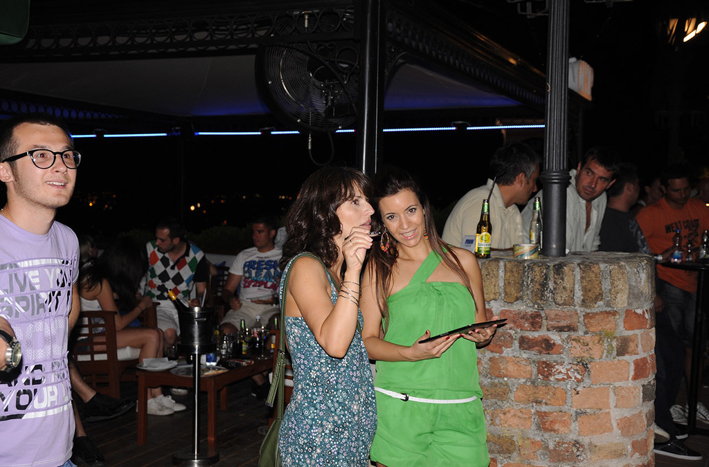 Two women stand close to each other at a social gathering, one talking on a mobile phone and the other holding a tablet. In the background, a rooftop bar scene with patrons and lit-up decor suggests a lively night out with interactive digital art elements.