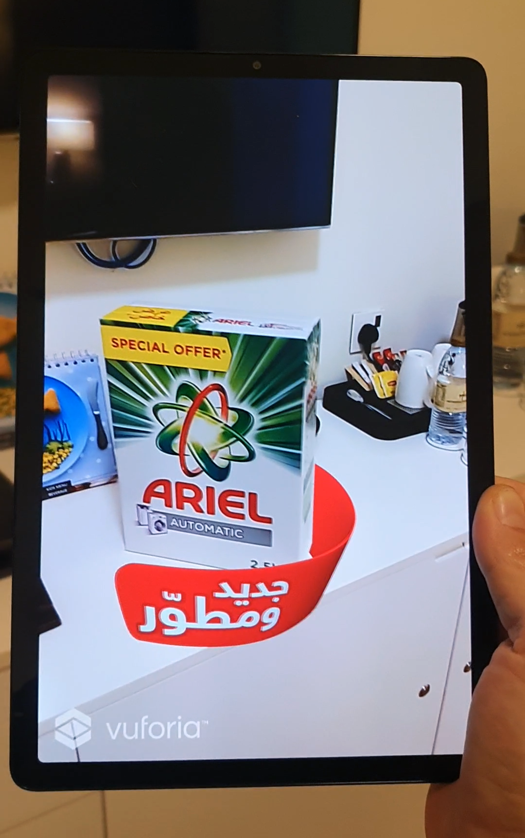 Close-up of a smartphone held in a hand, with the screen showing an augmented reality (AR) animation activated by scanning an Ariel laundry detergent box.