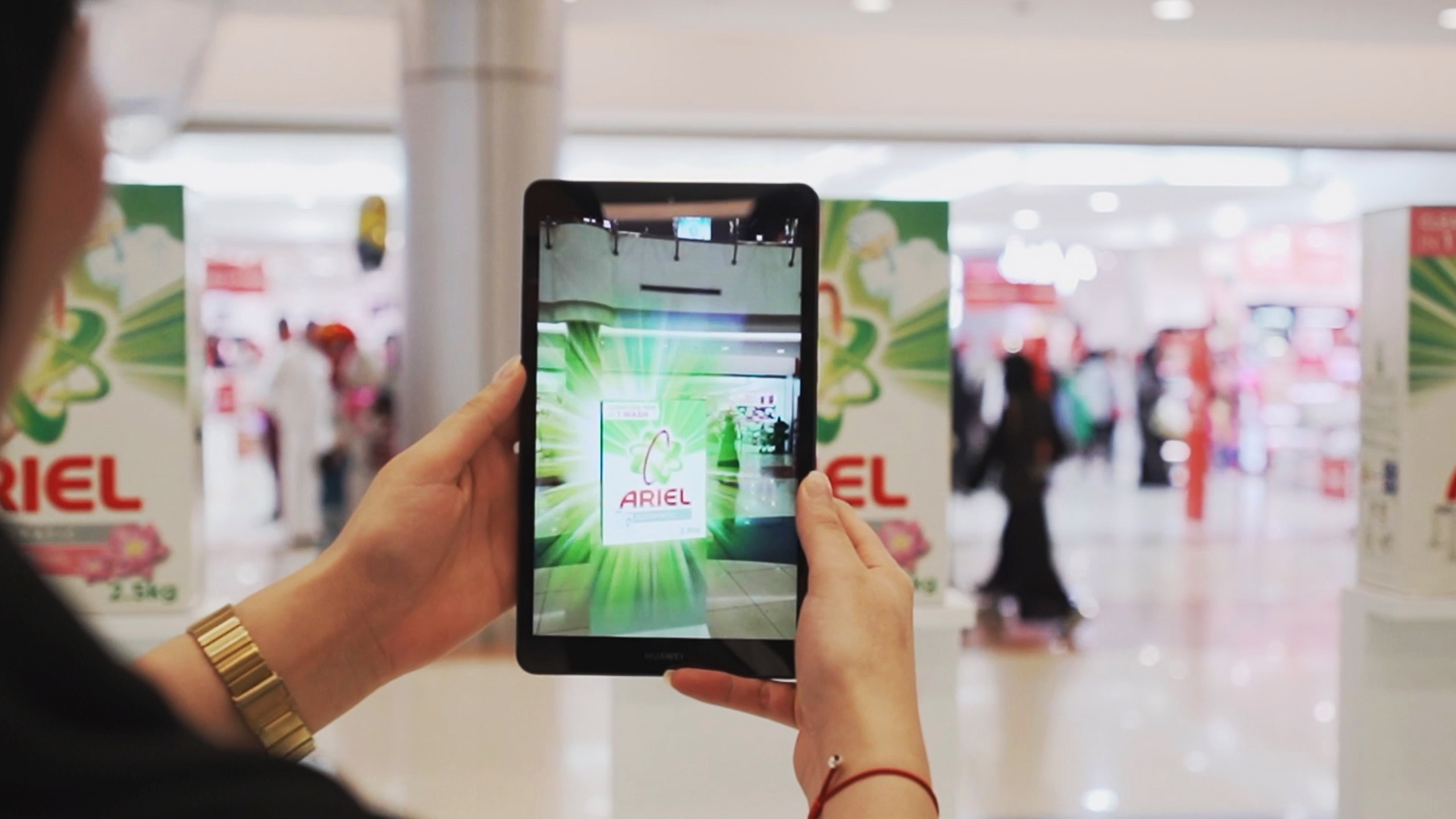 Over-the-shoulder view of a person holding a tablet that displays an augmented reality (AR) animation of the Ariel brand. The tablet camera is directed towards an Ariel advertisement in a shopping mall, where the AR feature overlays engaging graphics and product information onto the real-world image, creating an immersive promotional experience.