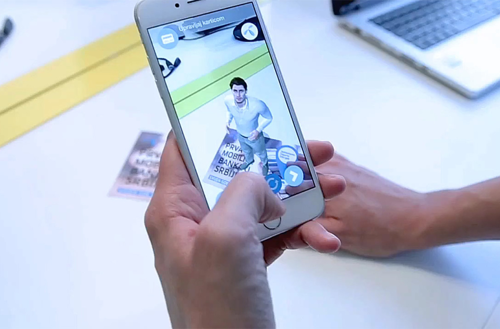 An augmented reality virtual banker is displayed on the screen of a smartphone held above a Telenor Bank card. The virtual figure, depicted as a friendly male character in a casual stance, introduces the mobile banking services of Telenor Bank in Serbia.