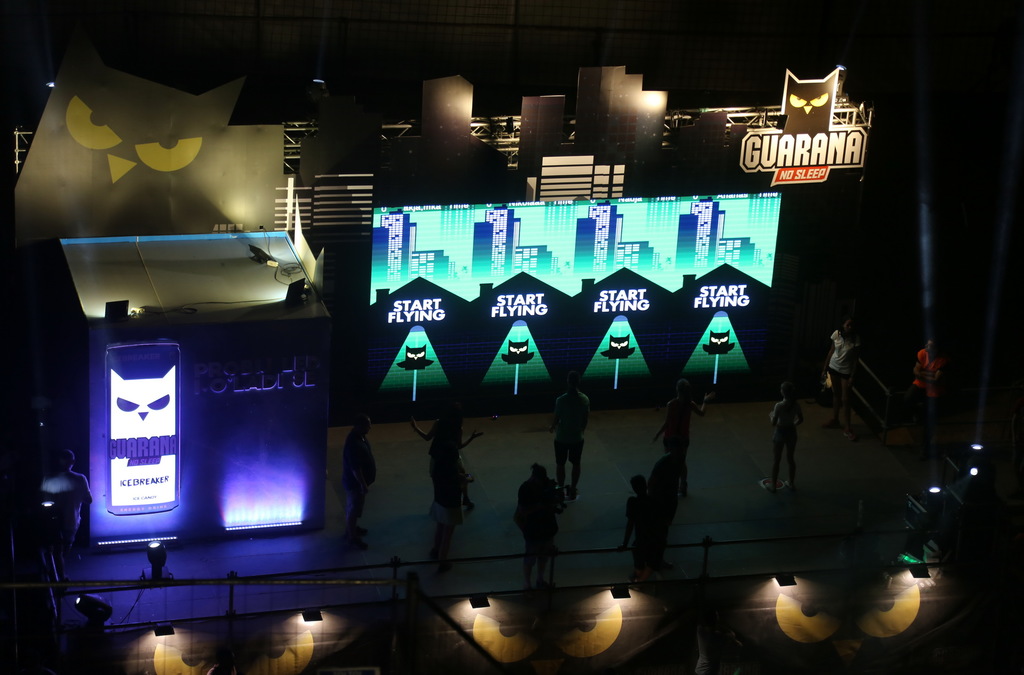 Overhead view of a darkened event space with a large LED wall displaying the Guarana logo and an interactive owl game, with participants and onlookers gathered around in a dimly lit atmosphere.