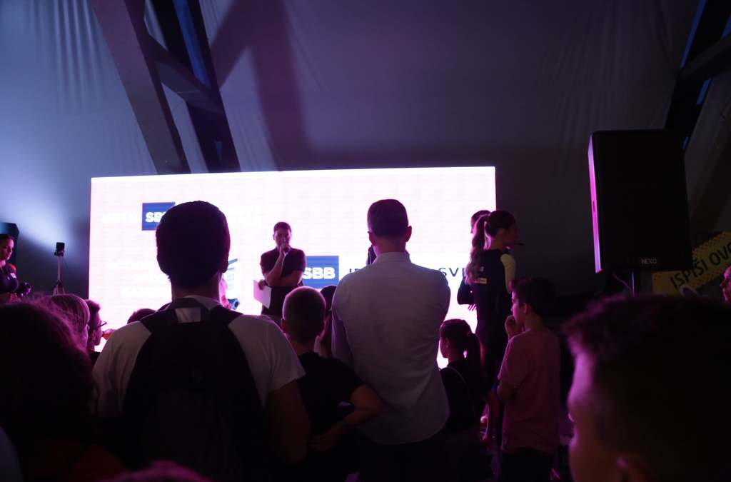 A silhouette view of a crowd gathered at the Balkan TUBE Fest around the SBB VR gaming booth, with a bright screen in the background. 