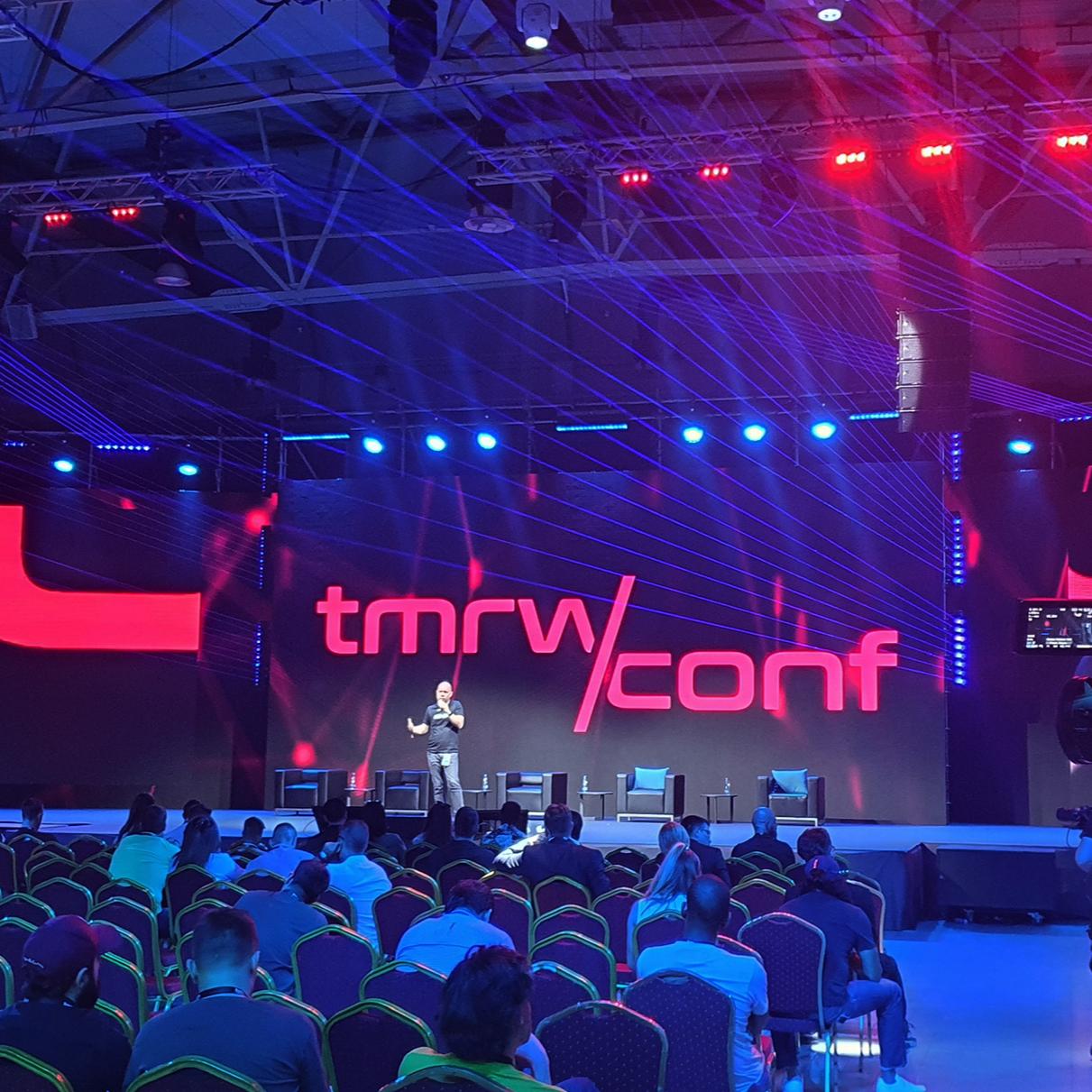 The TMRW Conference session in progress with a speaker on stage discussing the future of digital art, set against a dramatic backdrop of red and blue light beams, highlighting the event's focus on virtual reality and the arts.