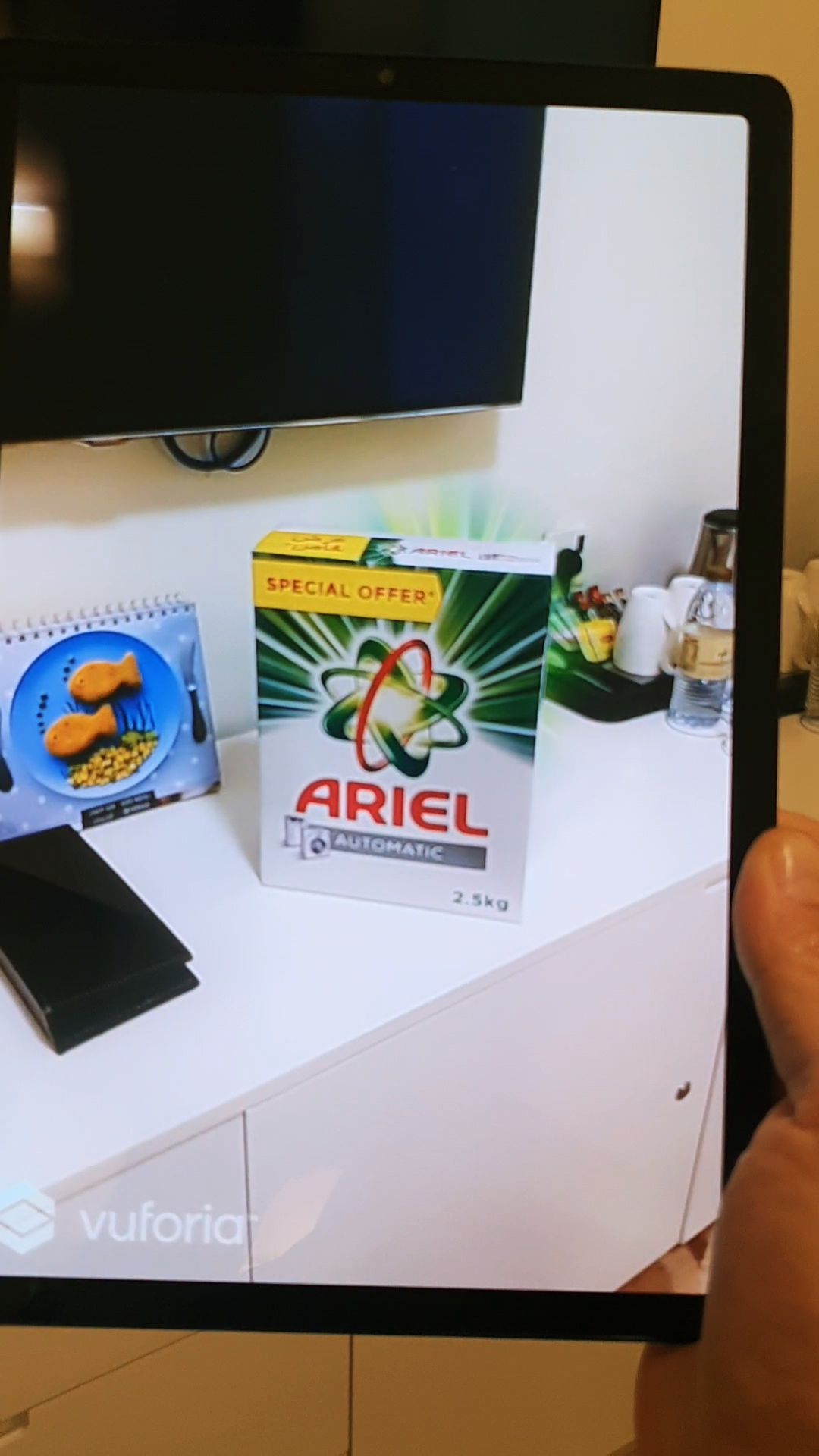 Close-up of a smartphone held in a hand, with the screen showing an augmented reality (AR) animation activated by scanning an Ariel laundry detergent box.