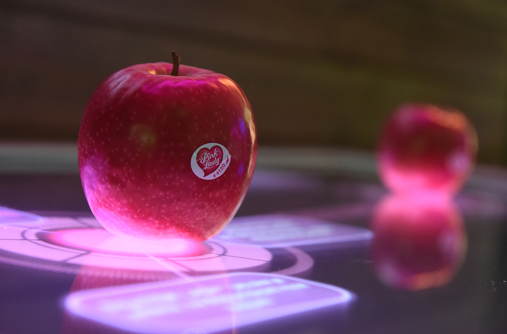 A red apple with a sticker sits atop a high-tech, illuminated multi-touch console displaying vibrant, interactive graphics around the apple, creating an engaging way to learn about the product in an exhibit.