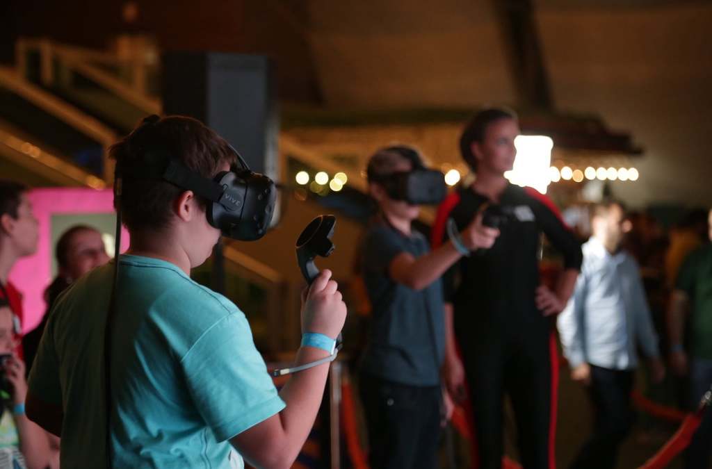 Two attendees at the Balkan TUBE Fest immersed in a VR experience, wearing headsets and holding controllers, with a crowd visible in the soft-focus background.