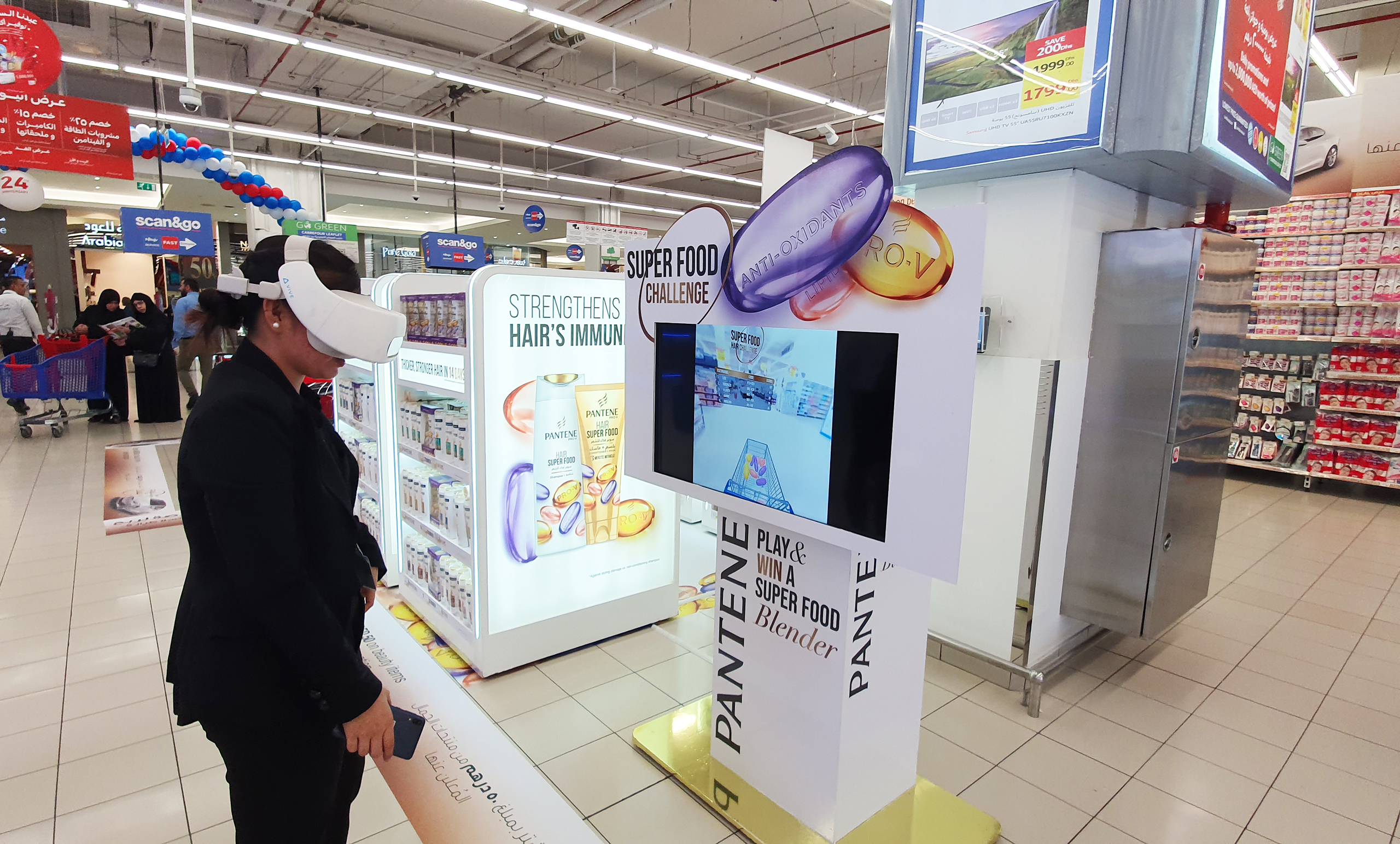 A person wearing a virtual reality headset is engaged in the 'Super Food Challenge' game at a promotional kiosk for Pantene's Hair Super Food shampoo. The kiosk is adorned with product branding and slogans about hair immunity boosted by vitamins. Shelves with boxed products are visible in the background.