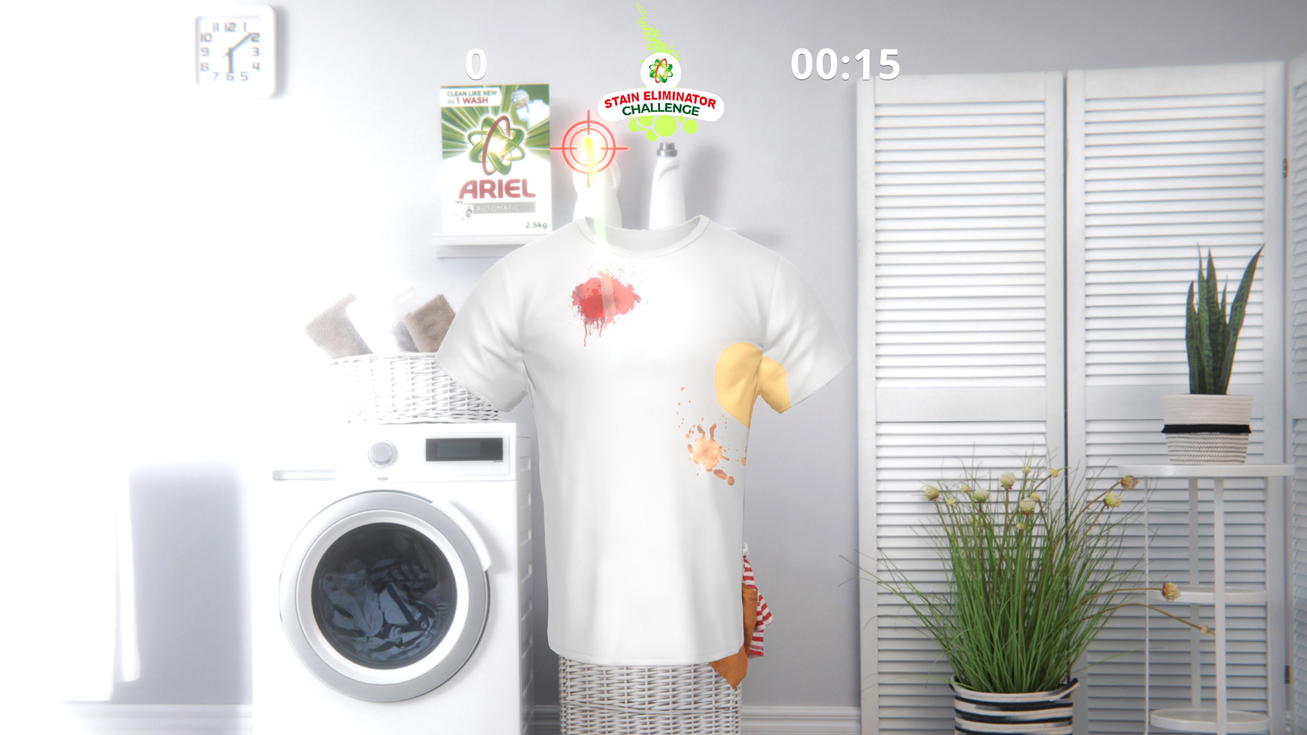 An engaging and bright virtual laundry setup displays the Ariel "Stain Eliminator" Challenge, a Kinect game. A white T-shirt displayed on a mannequin within a simulated laundry room shows a score of "0" and a timer set at "00:15". The shirt is stained with a red splash and a brown spill, with an Ariel product box visible on a shelf, promoting the stain-removing efficiency of Ariel laundry detergent through this interactive game.