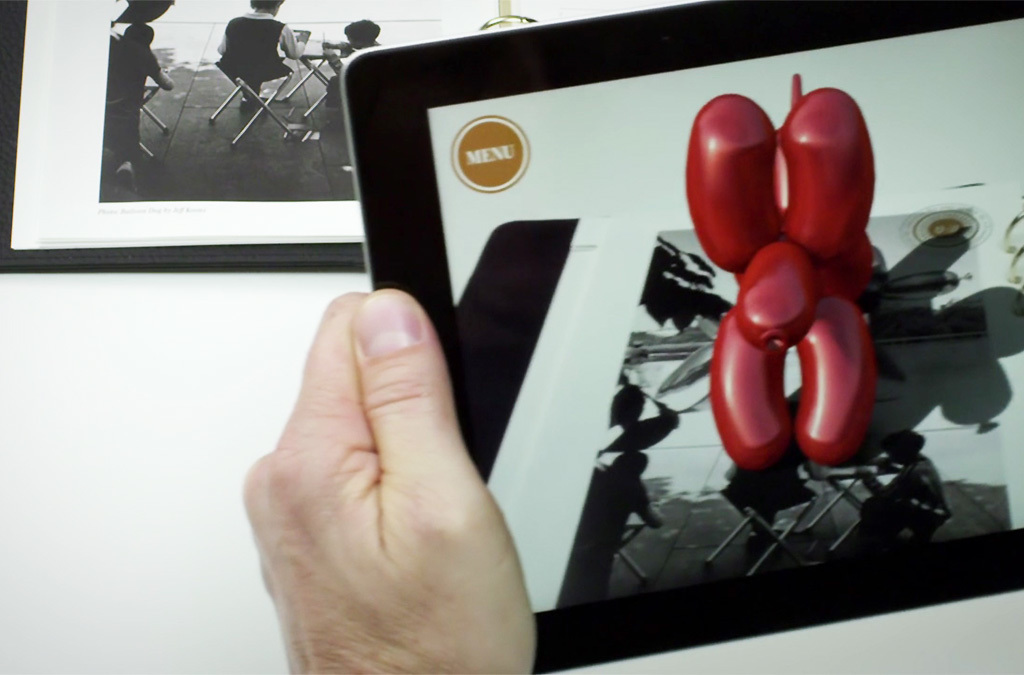 Hand holding a digital tablet over a page of the Signature Book, where the screen shows a 3D red balloon dog sculpture, exemplifying the augmented reality content unlocked by the Fifteen app.