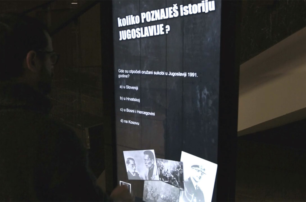 A museum visitor interacts with a question on a digital totem asking about the history of Yugoslavia, with multiple-choice answers displayed.
