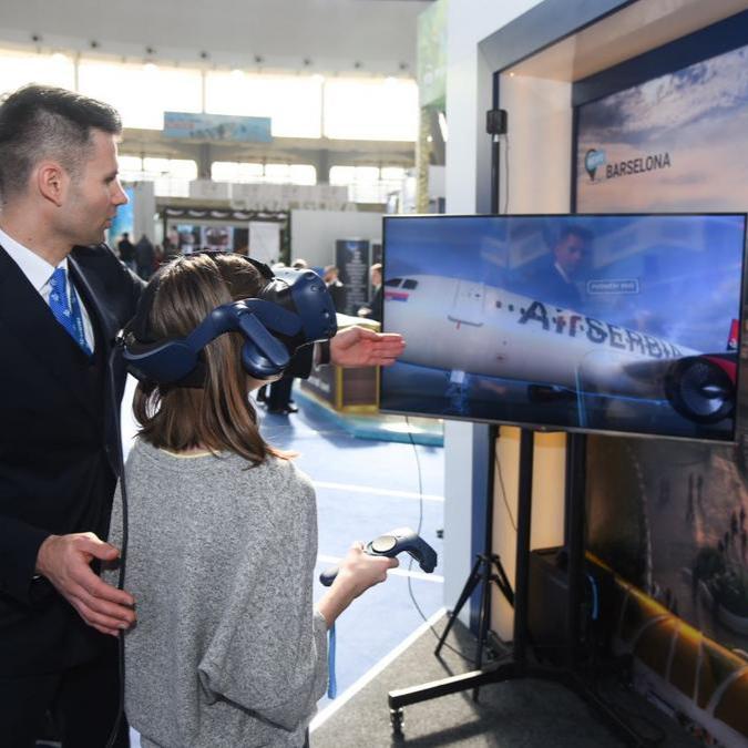 A woman wearing a VR headset is guided by an Air Serbia representative to explore a virtual aircraft.