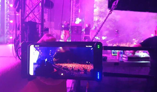 A creative perspective from behind a person's shoulder, holding a smartphone and recording a concert stage bathed in pinkish lights. The screen of the smartphone displays the crowd and stage, providing a framed view within the image. The surrounding stage elements are softly illuminated, enhancing the focus on the phone's screen.