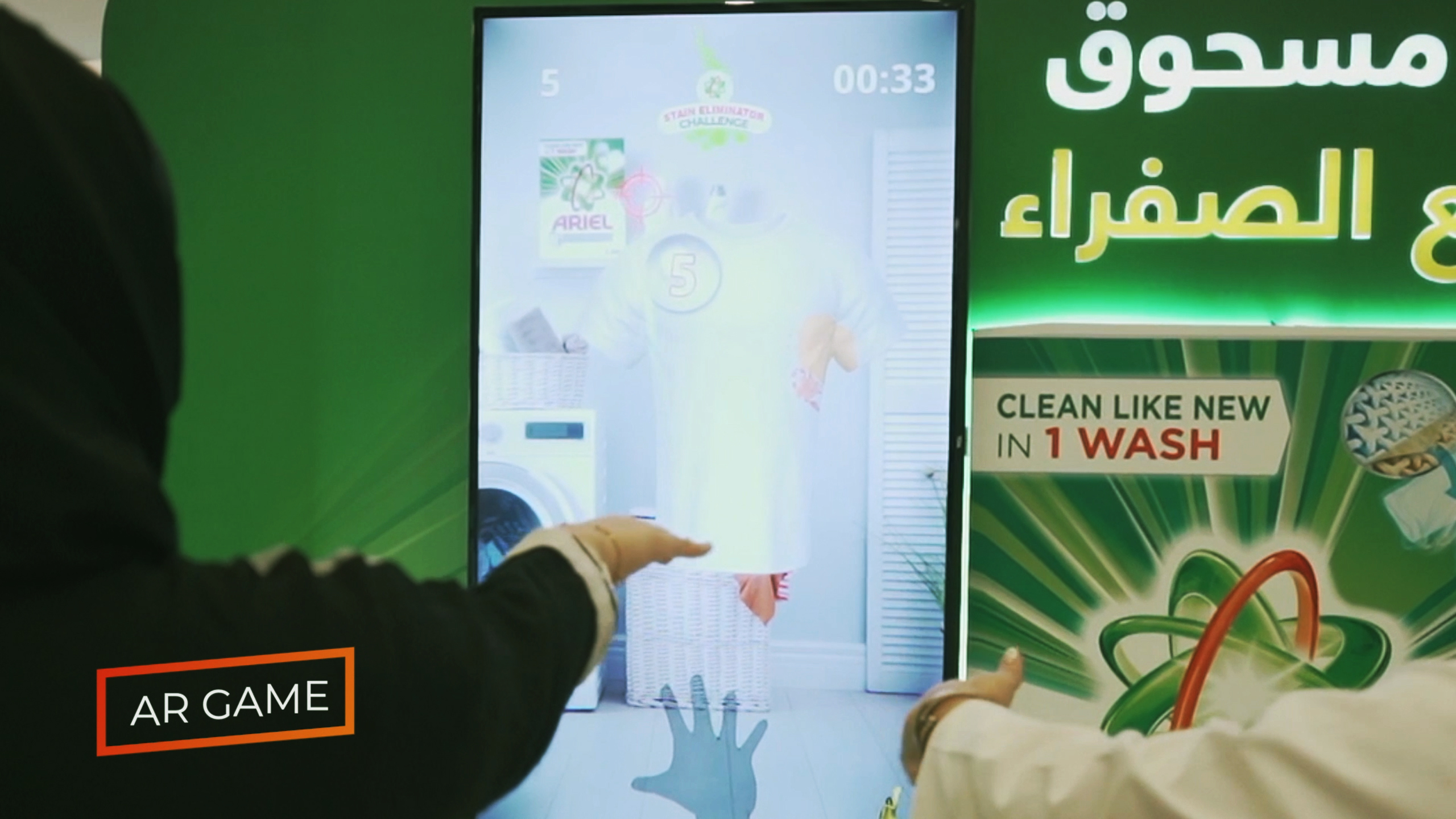 A person is playing the Ariel "Stain Eliminator" Kinect game, shown on a large screen that blends a physical gaming area with an augmented reality (AR) experience.