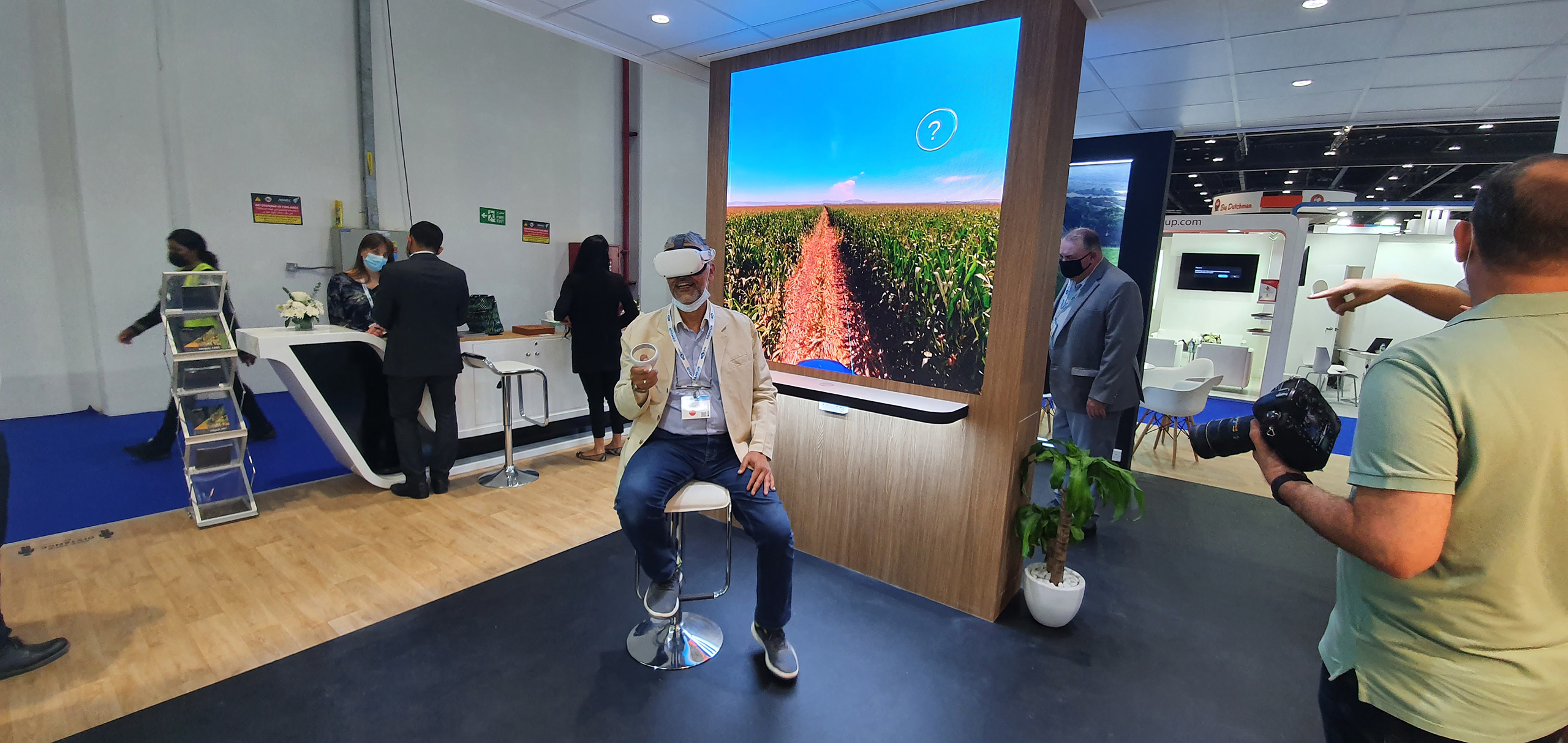 A man sits on a swivel chair wearing a virtual reality headset in a busy exhibition space, experiencing a VR simulation.