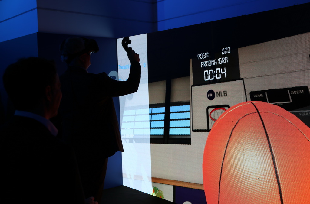 A participant with a VR headset is shown in mid-action, simulating a basketball shot toward a large screen displaying a virtual basketball hoop and score.
