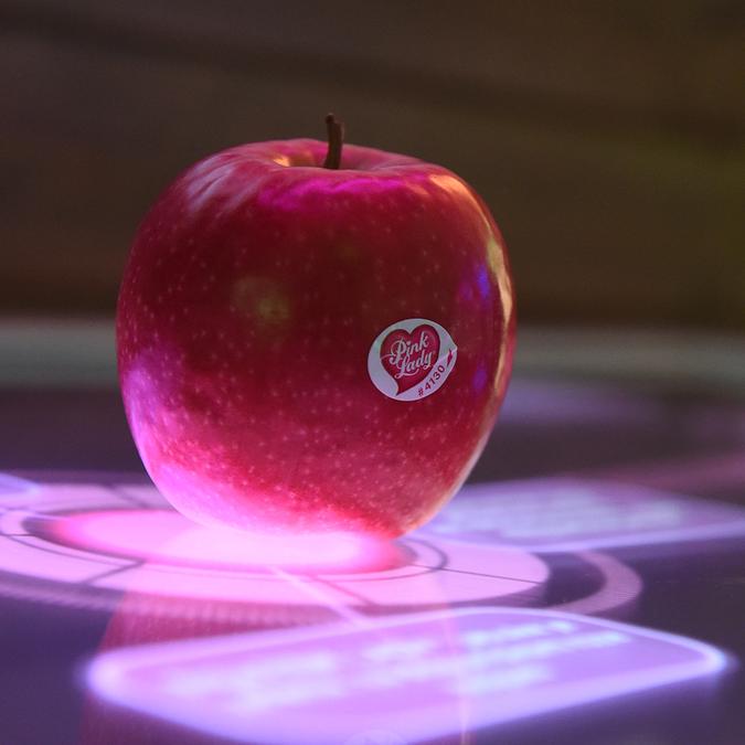 A red apple with a sticker sits atop a high-tech, illuminated multi-touch console displaying vibrant, interactive graphics around the apple, creating an engaging way to learn about the product in an exhibit.