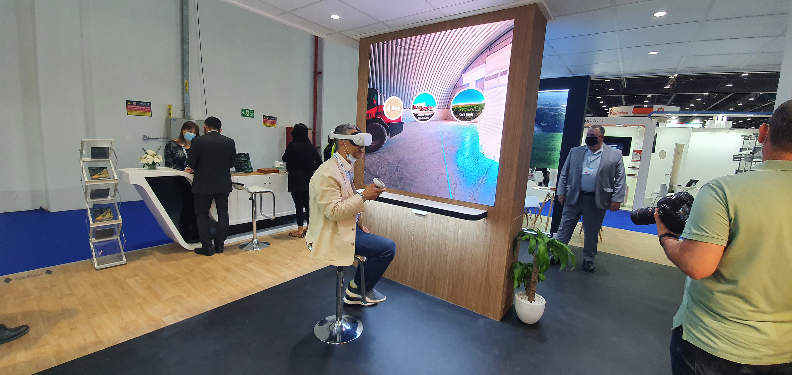  A man sits on a swivel chair wearing a virtual reality headset in a busy exhibition space, experiencing a VR simulation. 