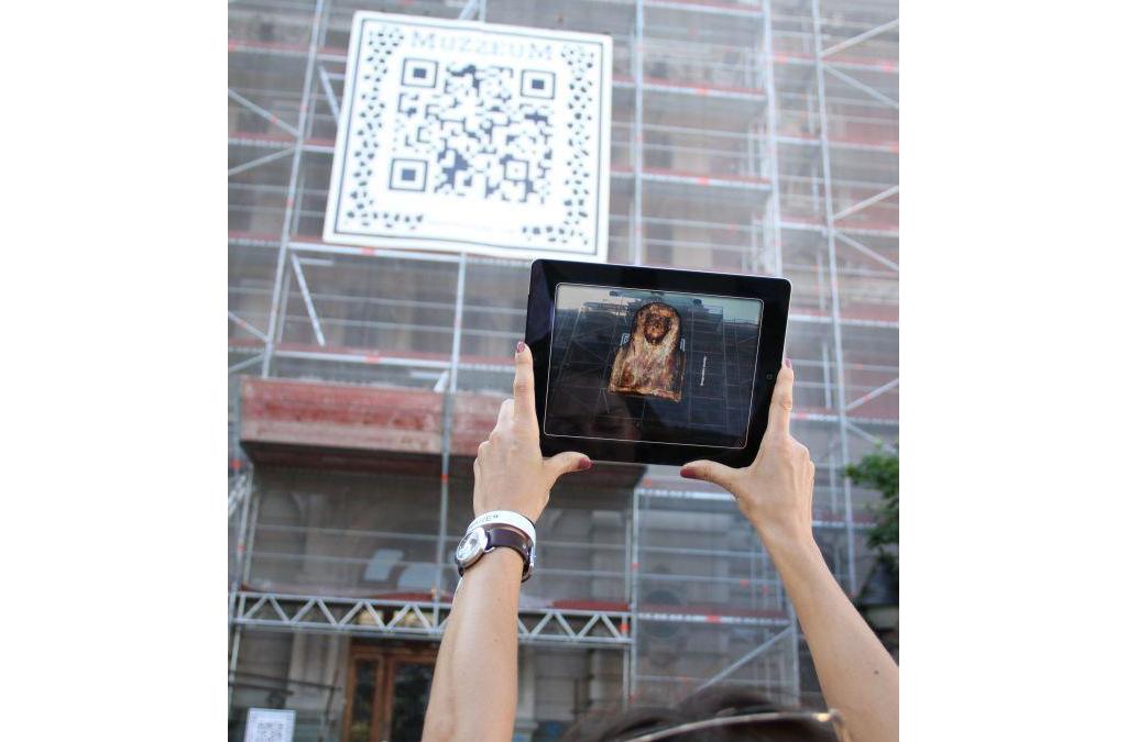A person uses a tablet to scan a special ARQR code from the MUZZEUM project displayed on a building, revealing a virtual artifact on their screen.