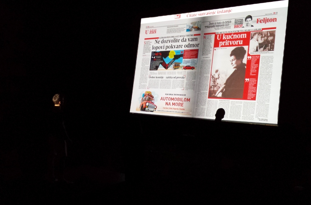 A person in a dark room using hand gestures to interact with a large projection of a digital newspaper, powered by Gesture Page Flipping software with a Microsoft Kinect sensor.