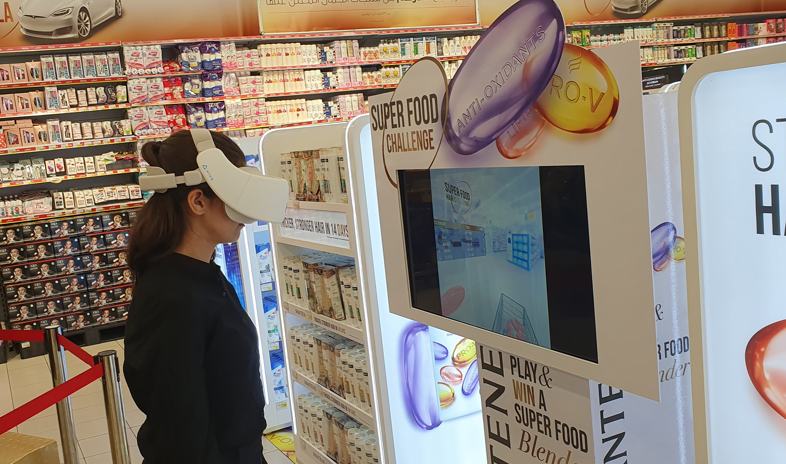 A person wearing a virtual reality headset is engaged in the 'Super Food Challenge' game at a promotional kiosk for Pantene's Hair Super Food shampoo. The kiosk is adorned with product branding and slogans about hair immunity boosted by vitamins. Shelves with boxed products are visible in the background.