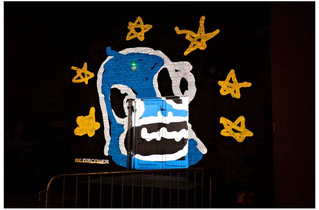A projected blue and yellow image of a screaming mouth surrounded by stars, created with laser painting, decorates a wall.