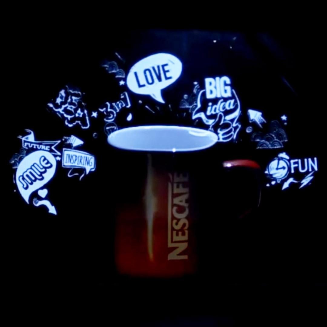 A Nescafe cup placed within a hologram pyramid, with animated words like "LOVE," "BIG IDEA," "SMILE," and "FUN" floating above it against a dark background, showcasing a blend of physical and digital display.