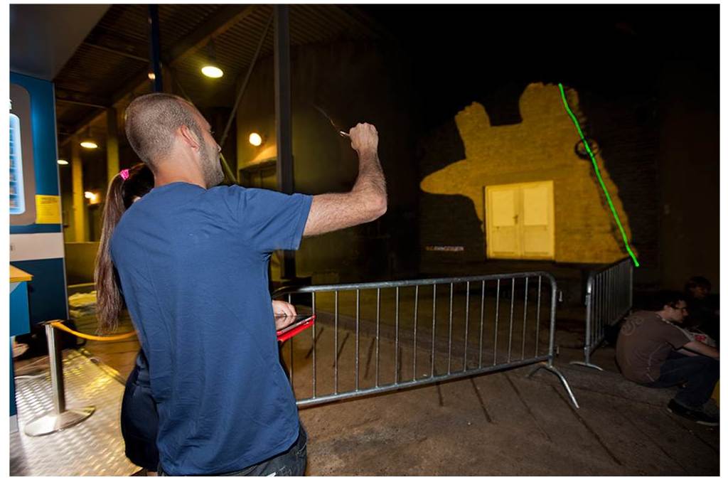 An individual is seen from behind, actively using a laser pointer to paint on a large surface in a dark setting, demonstrating the interactive nature of laser painting, where movements are captured by a special camera and translated into digital artwork.