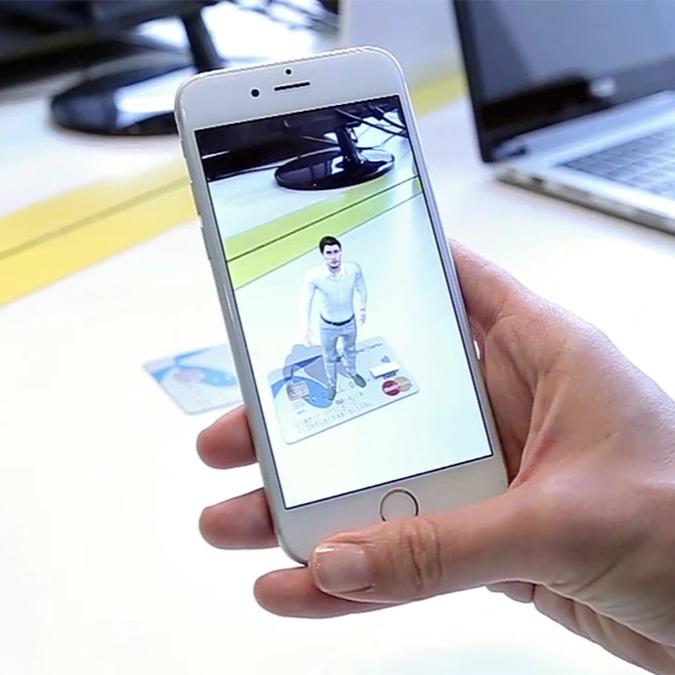 A person's hand holding a smartphone displaying an augmented reality (AR) image of a digital banker standing on a Telenor Bank card. The digital banker is gesturing welcomingly within the screen of the smartphone, which is being viewed over a desk with a laptop in the background.