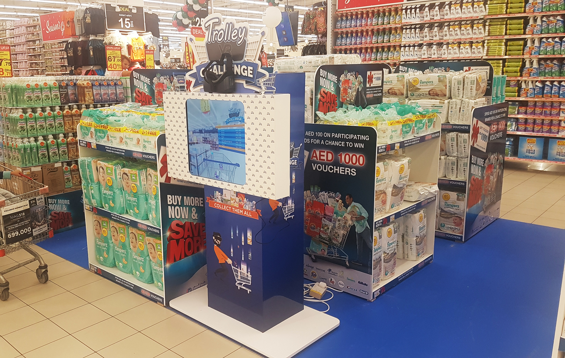 A promotional stand for 'VR Trolley Challenge' situated in a bustling Carrefour store. The stand features vibrant signage and a screen showing a virtual shopping basket. Products and special offers are displayed around the area, with customers, including a young child in a shopping cart, browsing nearby.