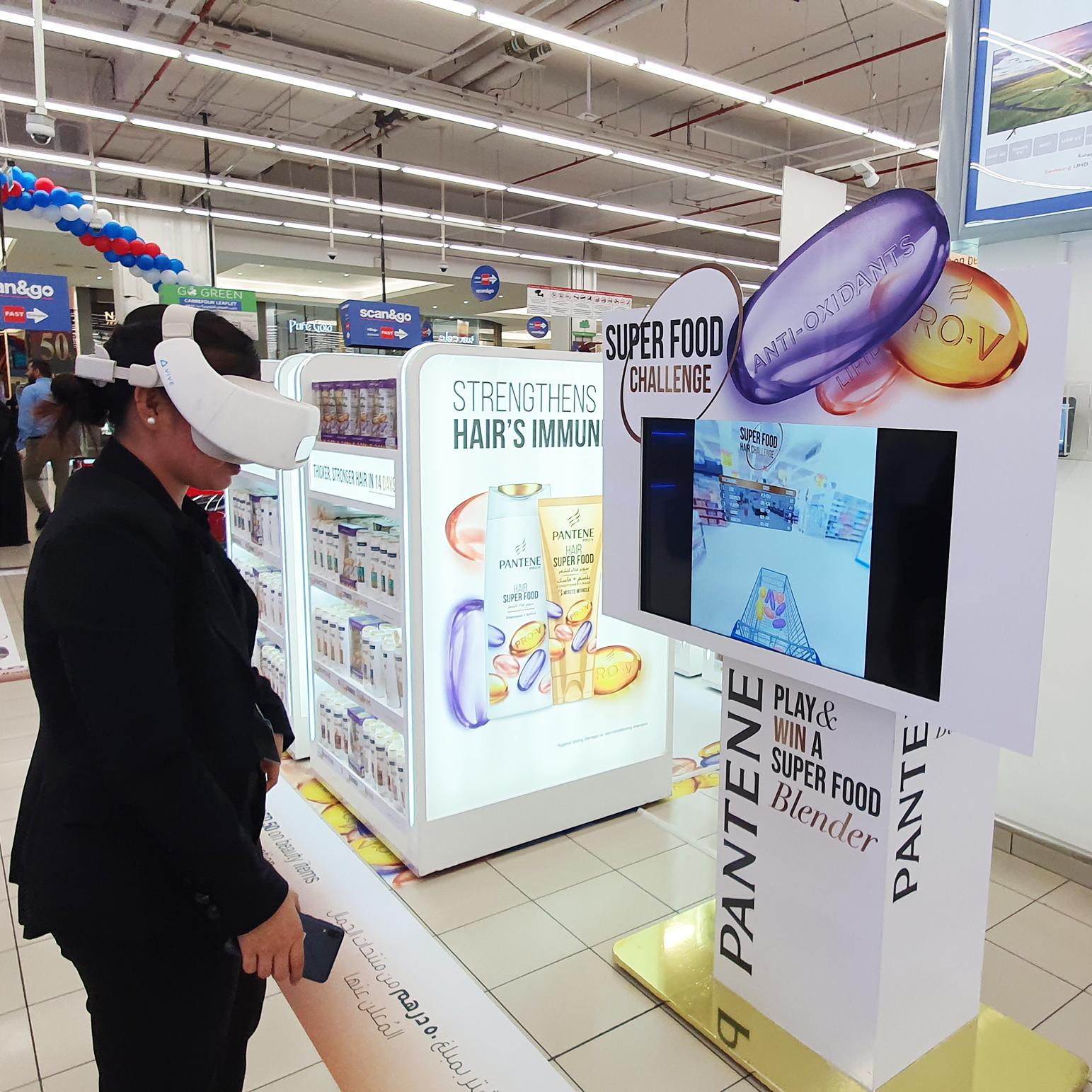 A person wearing a virtual reality headset is engaged in the 'Super Food Challenge' game at a promotional kiosk for Pantene's Hair Super Food shampoo. The kiosk is adorned with product branding and slogans about hair immunity boosted by vitamins. Shelves with boxed products are visible in the background.