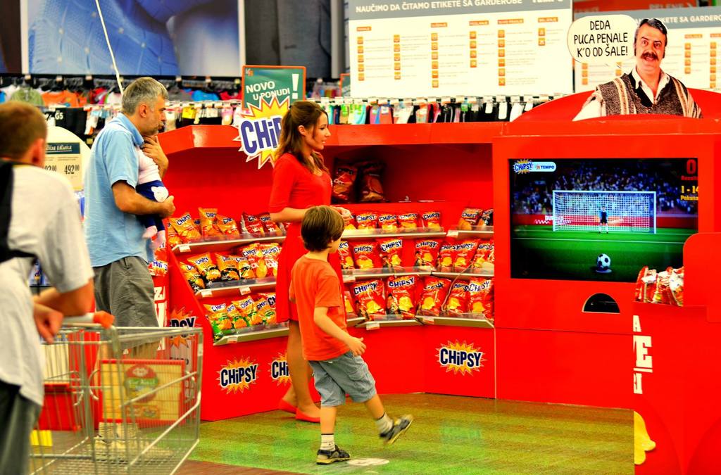 Shoppers in a grocery store pass by a promotional setup for Chipsy, featuring a soccer penalty kick interactive game, blending retail with entertainment.
