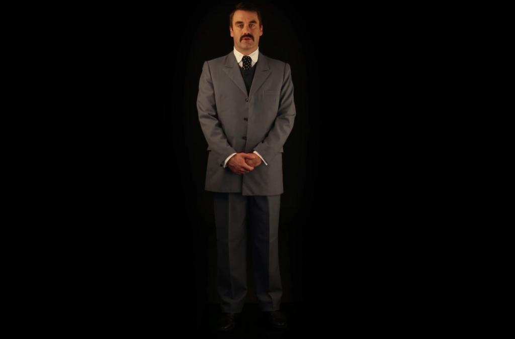 A man in a suit standing against a black background.