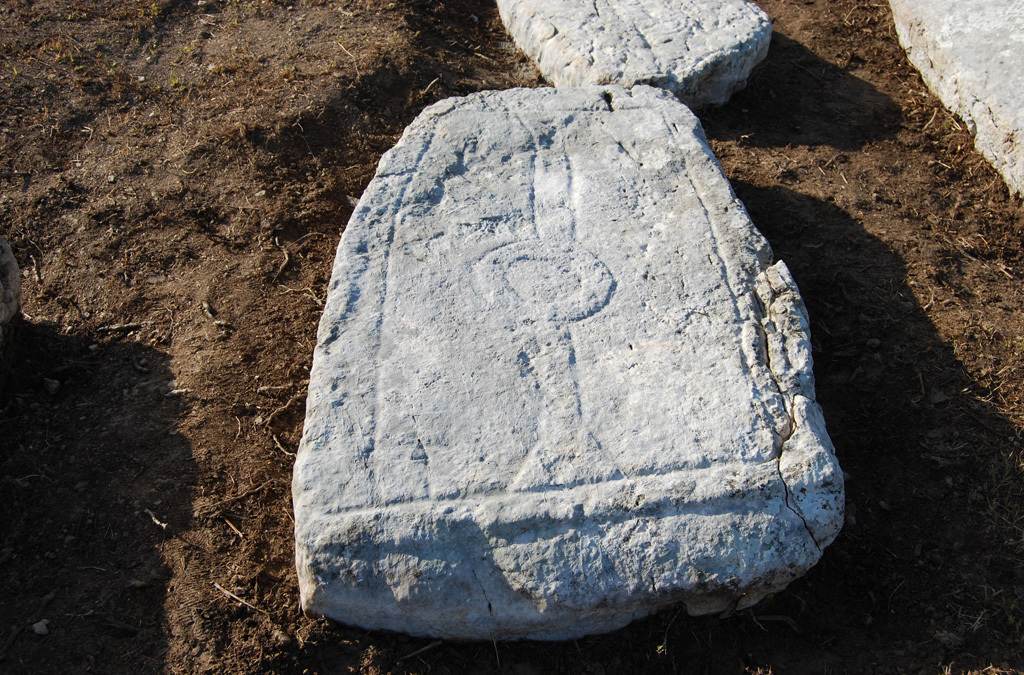 Image of an ancient tombstone inscribed with symbolic patterns, situated on the ground at the archaeological site.