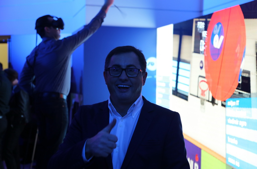 Smiling man giving a thumbs up in the foreground with another man wearing a VR headset and interacting with the game in the background.