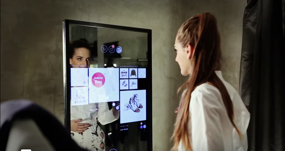 A woman interacts with a high-tech mirror in a fitting room, displaying various clothing items and virtual buttons for a premium shopping experience.