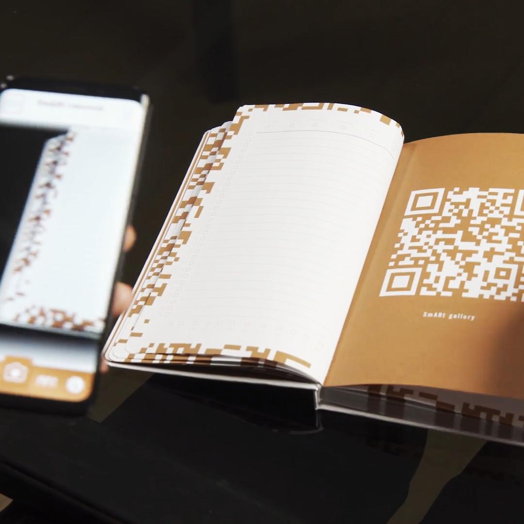 A user's hand holds a smartphone with the SmARt AR app, capturing a QR code on an open Banca Intesa planner to convert written content into a digital entry.